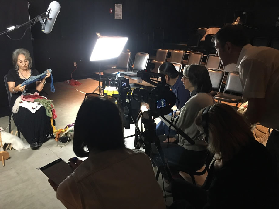 A group of people stand behind a camera filming while a woman sits under a light being filmed.