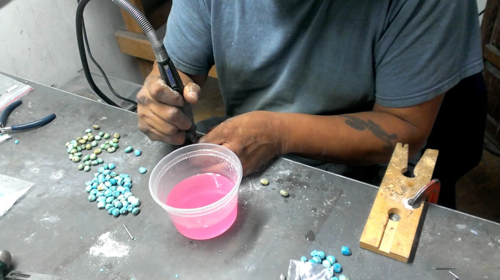 A jeweler's hands hold a tool atop a workbench with turquoise stones. 